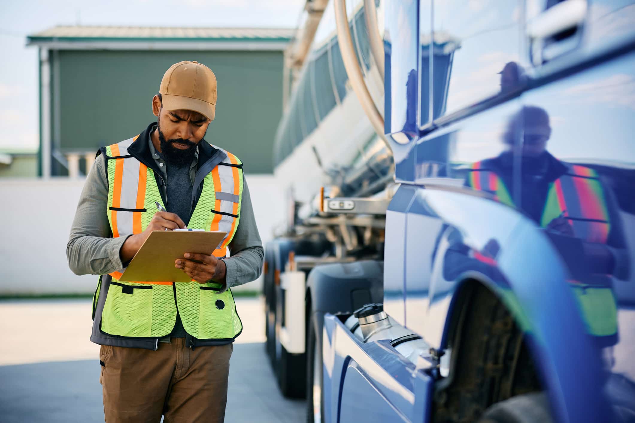A CNG technician wearing a hi-vis vest and cap holds a clipboard while performing a pre-service inspection on a heavy-duty vehicle's CNG fuel system.